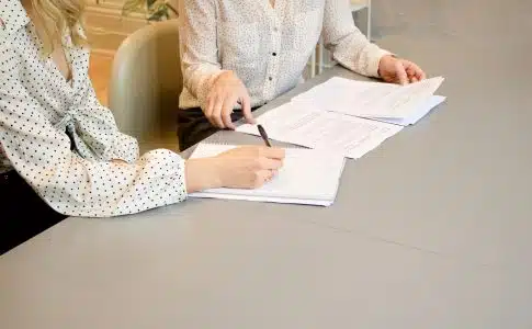 woman signing on white printer paper beside woman about to touch the documents